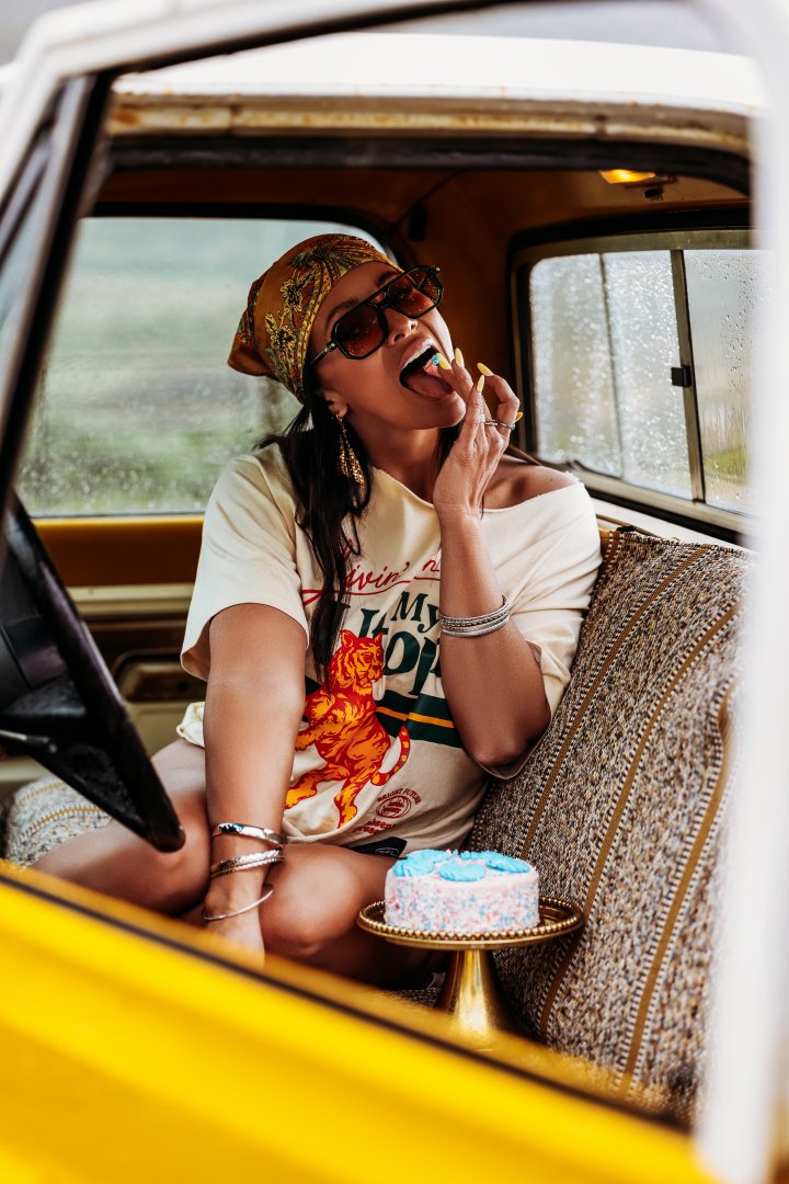 Woman sitting in a vintage vehicle, wearing sunglasses and a patterned headscarf, eating a slice of cake, with a small birthday cake on a tray in front of her, on a rainy day.