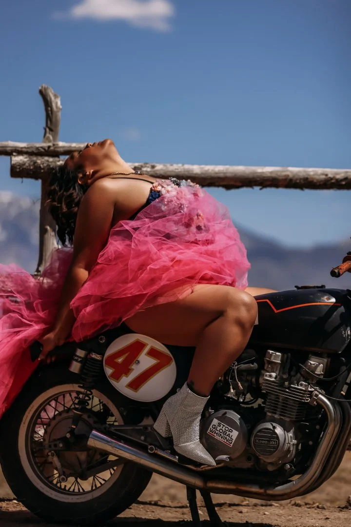 A woman in a pink tulle dress is sitting on a black motorcycle, with her head tilted back and eyes closed, against a background of a blue sky with some clouds and a wooden fence.