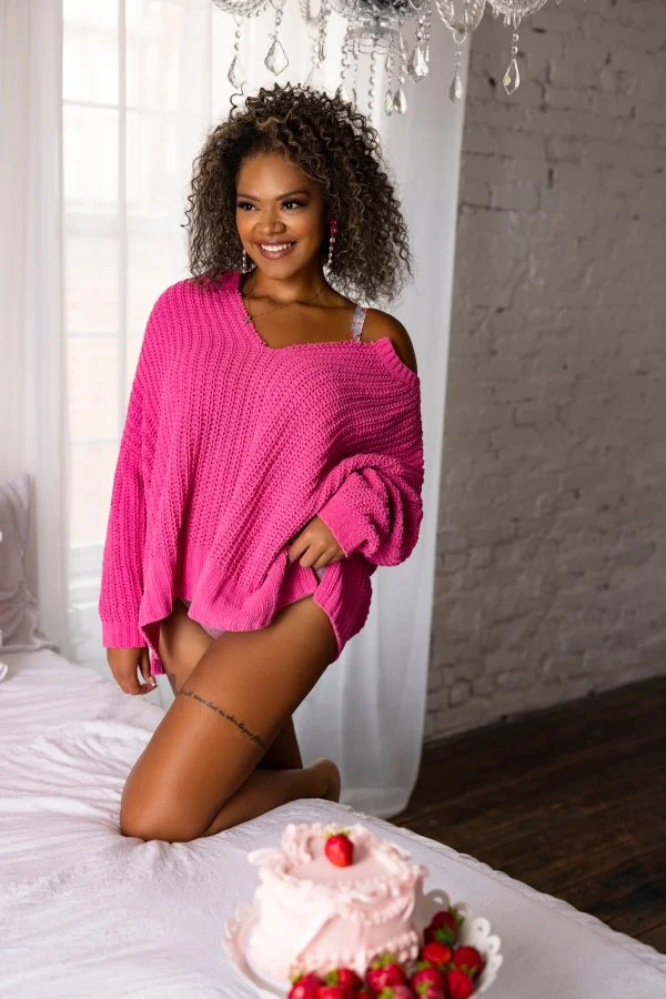 A young woman with curly hair wearing a pink sweater and matching pink shorts, kneeling on a bed with a bakery cake and strawberries in front of her, in a light-filled room with a chandelier and brick wall.