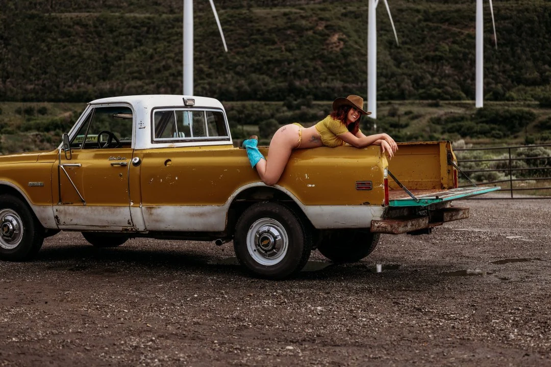 A woman in a yellow top, hat, and blue socks, lying in the truck bed of a vintage yellow and white pickup truck with a mountain landscape and wind turbines in the background.
