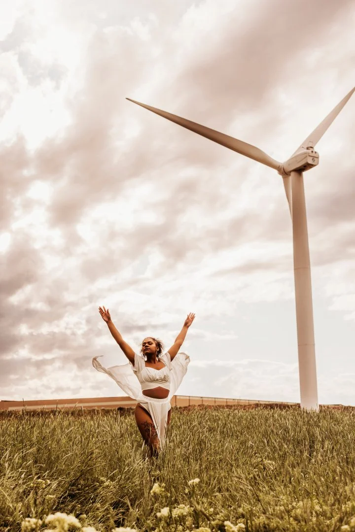 A woman in a white dress with open arms stands in a grassy field near a large wind turbine on a cloudy day.