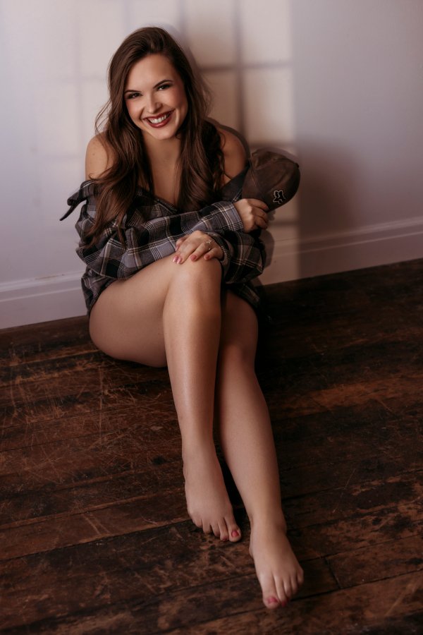 A young woman with long brown hair sitting on a dark wooden floor, smiling and holding a cap, wearing a plaid shirt and no pants.