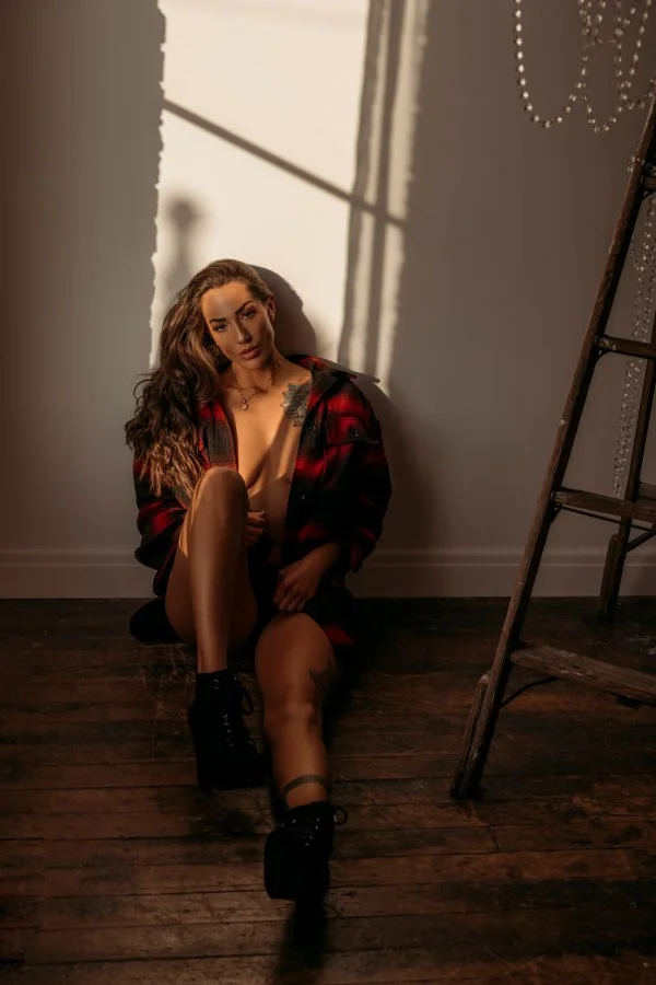 Woman sitting on a wooden floor in a sunlit room, wearing a red and black plaid shirt, black boots, and fishnet stockings, with a shadow cast on the wall behind her.
