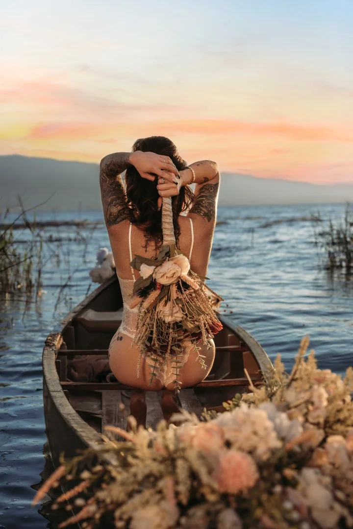 A woman with tattoos sitting in a boat on a lake during sunset, holding a decorative bouquet behind her back, with flowers in the foreground.