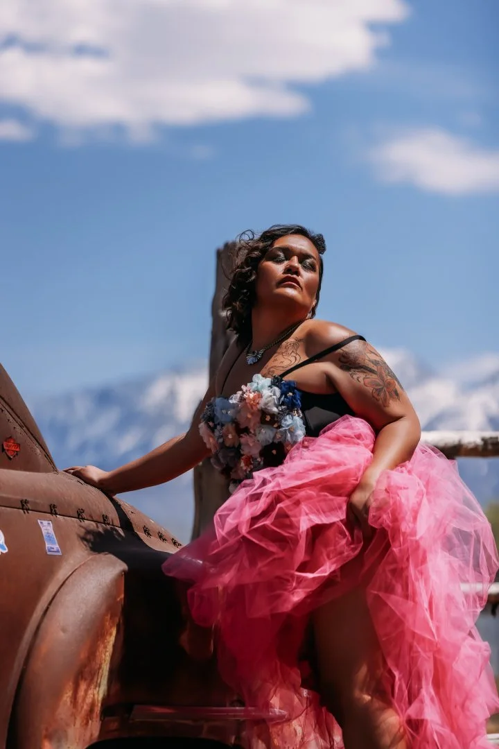A woman in a pink tulle skirt and black top with floral embellishments, with tattoos on her arm, posing on a rusted vintage vehicle outdoors against a mountain landscape and cloudy sky.