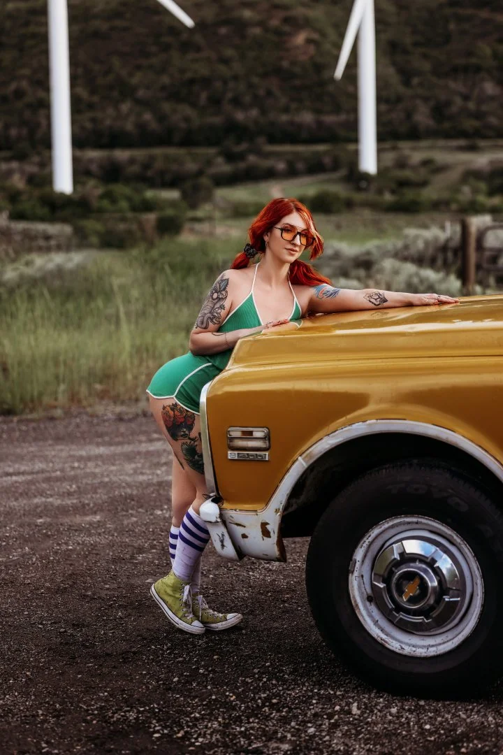 A woman with red hair, tattoos, and sunglasses leaning on the front of a yellow vintage car, dressed in a green outfit, with wind turbines in the background.