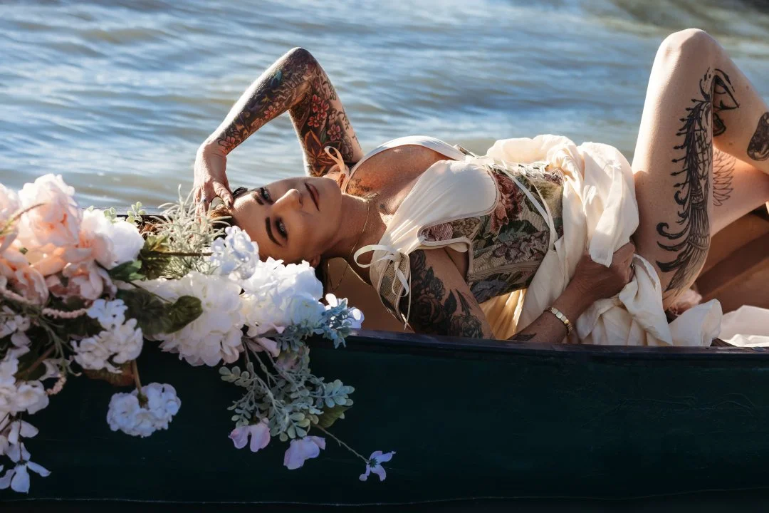 A woman with tattoos lying in a boat on the water, surrounded by flowers, wearing a vintage dress.