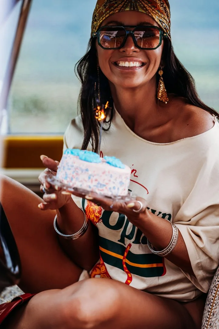 Woman smiling and holding a birthday cake with blue decorations, wearing sunglasses, a colorful headscarf, and earrings, enjoying a moment at the beach.