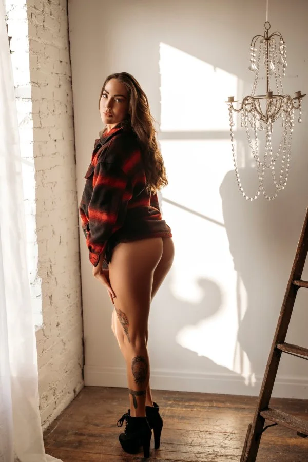 A woman standing near a window with sunlight casting shadows, wearing a plaid shirt and high heels, with tattoos visible on her leg.