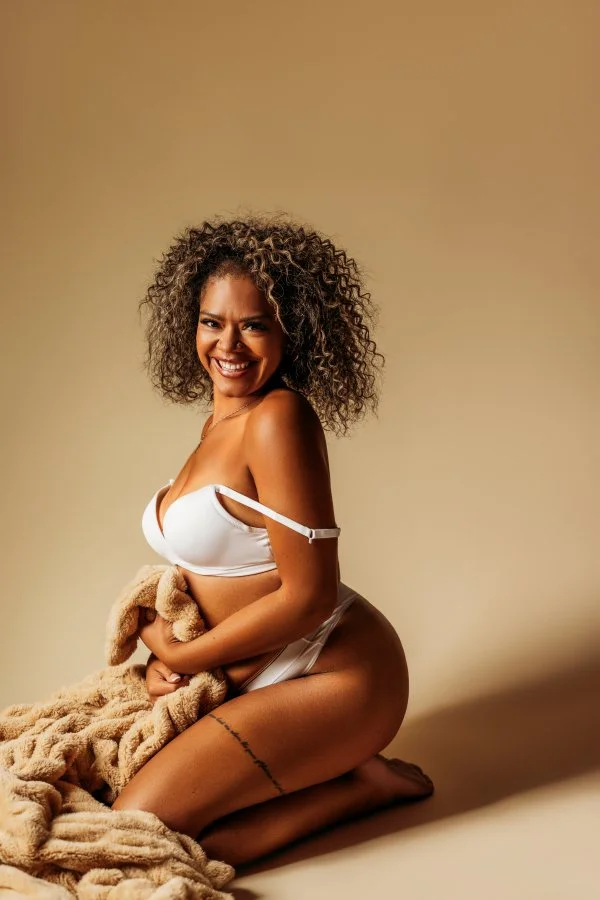 A woman with curly hair smiling and kneeling on the floor, wearing white lingerie and holding a beige blanket.