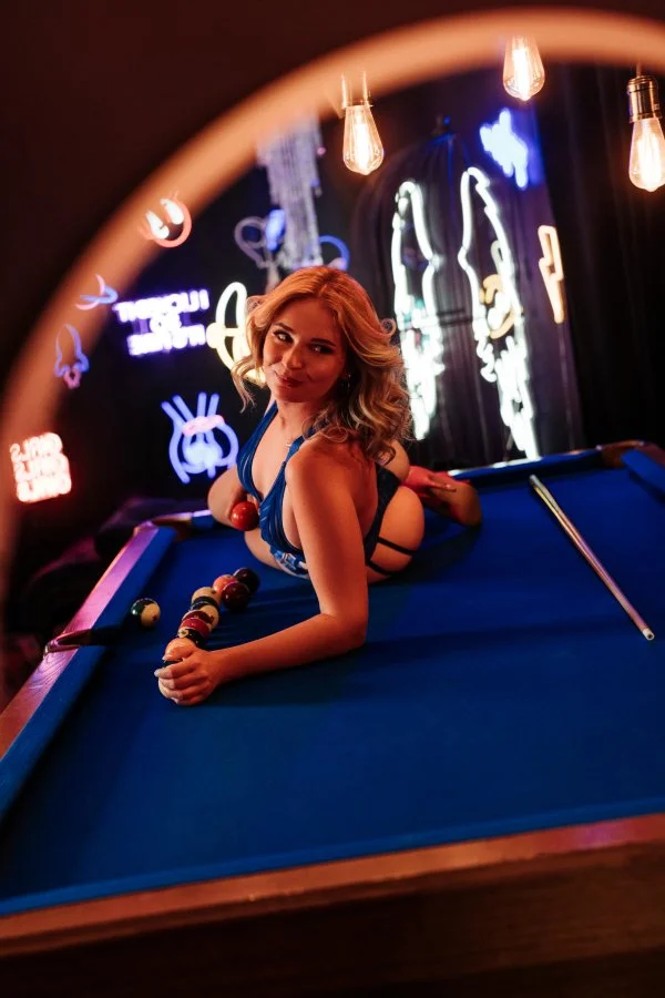 A woman in a blue outfit is lying on a pool table, holding pool balls, with neon lights and hanging bulbs in a dimly lit room.