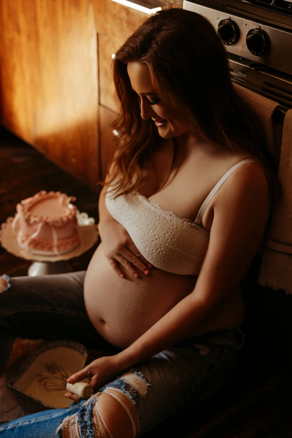 A pregnant woman smiling in a cozy kitchen, hands resting on her belly beside a decorative cake