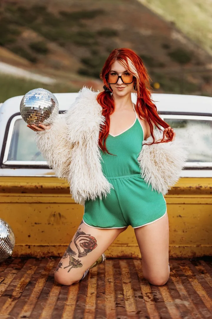 A woman with red hair, glasses, and tattoos, wearing a green romper and a white furry jacket, holding a disco ball, standing in front of a vintage yellow truck with mountains in the background.