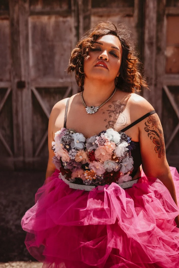 Woman wearing a floral patchwork top with a bright pink tulle skirt, posing outdoors in front of a wooden barn with a confident expression.