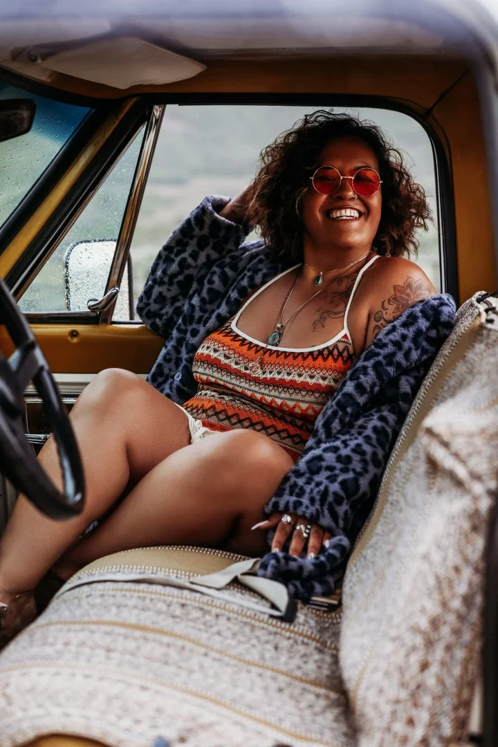 A woman sitting in a vintage car, smiling and wearing sunglasses, a colorful patterned tank top, a blue leopard print jacket, and jewelry, with wet weather visible outside.