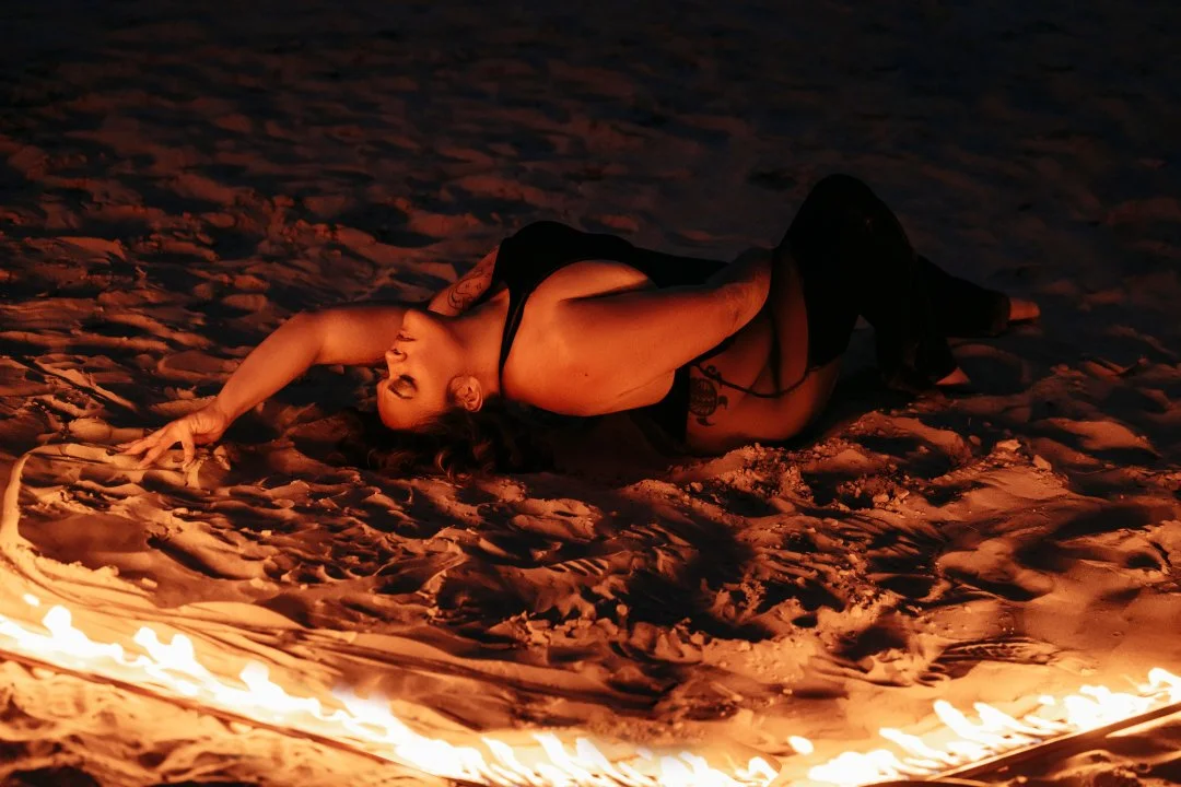 Person lying on the sand near a ring of fire at night, posing with one arm extended and head tilted back, dressed in black with tattoos visible on their body.