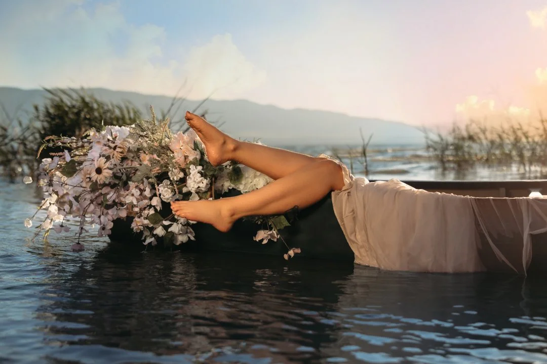 Person lying in a boat surrounded by flowers in water with a sunset and mountain background.