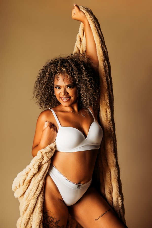 A woman with curly hair smiling while wearing white lingerie and holding a beige fur coat against a plain background.