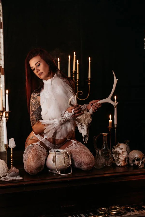 Kneeling fallen angel in lace and feathers with deer skull on piano, dark Halloween boudoir photography
