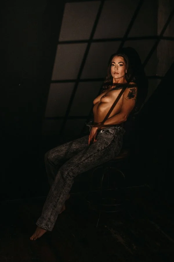 A topless woman with dark hair and a tattoo sitting on a high stool in a dark room, illuminated by soft lighting, with a grid-patterned ceiling in the background.