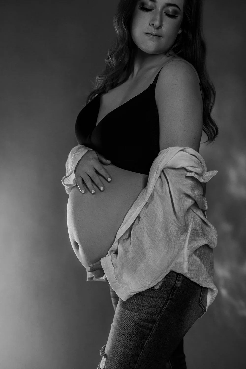 A black-and-white portrait of a woman with a shirt off the shoulders, hand resting on her abdomen