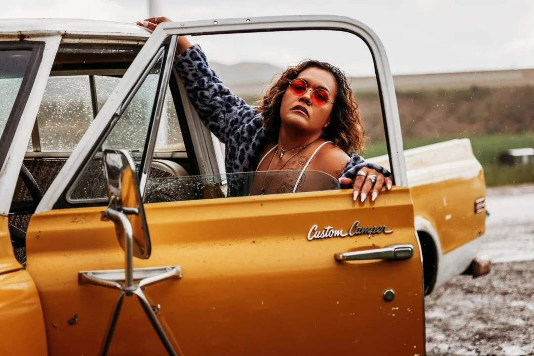 A woman with curly hair, wearing sunglasses, a leopard print jacket, and a tank top, leaning out of a vintage yellow truck labeled 'Custom Camper', in a rural outdoor setting with cloudy skies and wet ground.