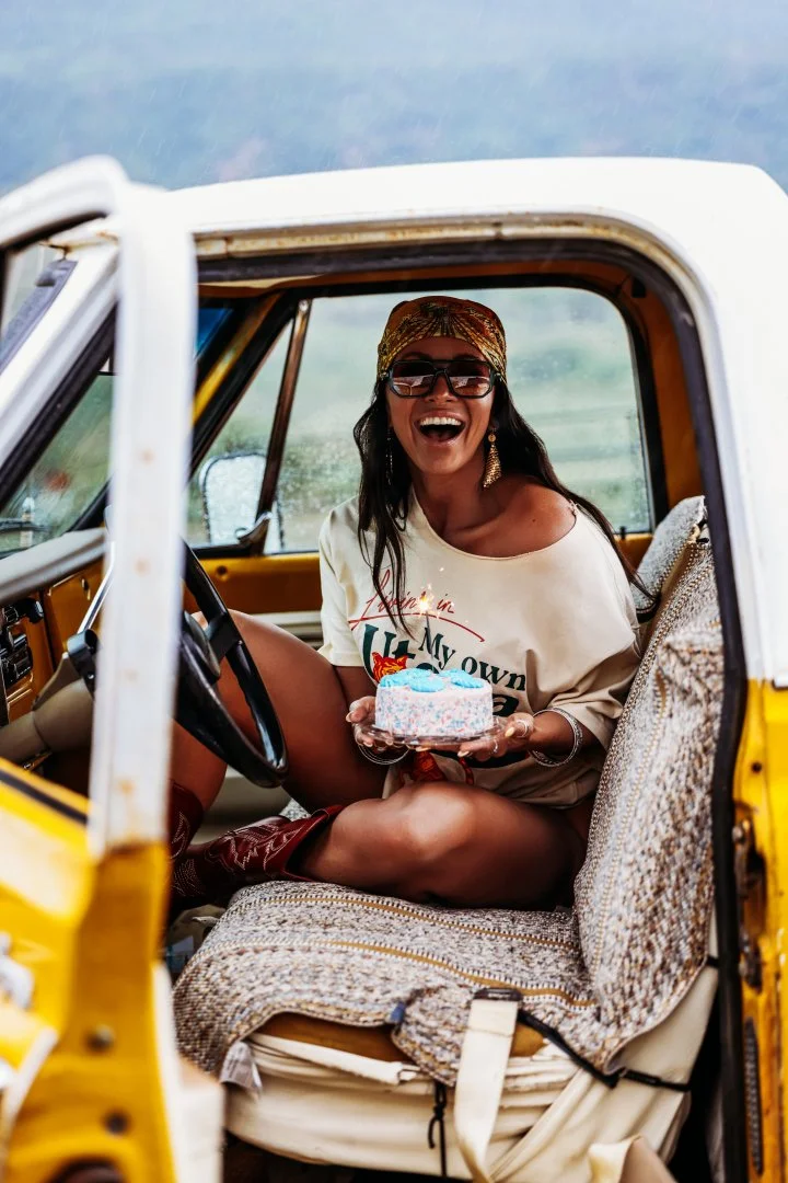 Woman sitting inside a yellow vintage car, smiling, holding a small birthday cake, wearing sunglasses, a headscarf, and casual clothes, with a scenic landscape in the background.