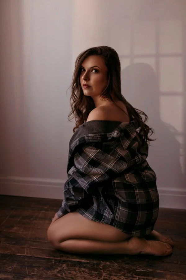A woman with long wavy hair sitting on the wooden floor, wearing an oversized plaid shirt.
