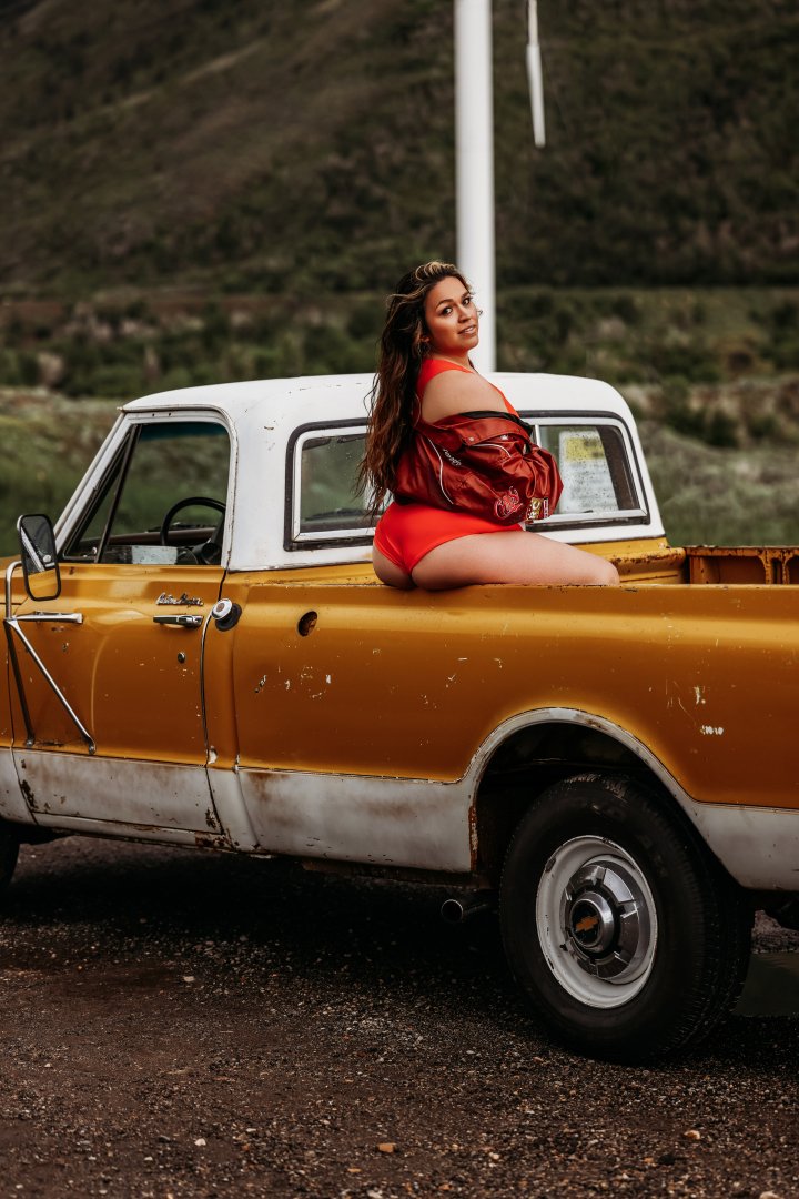 A woman in a red dress and jacket sitting in the bed of an old yellow and white pickup truck parked outdoors with a rural landscape in the background.