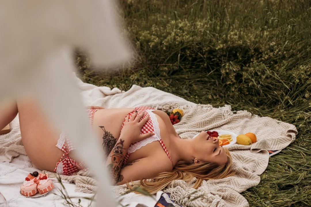 A woman lying on her back on a blanket outdoors, wearing a red and white checkered bikini, with food and flowers around her.