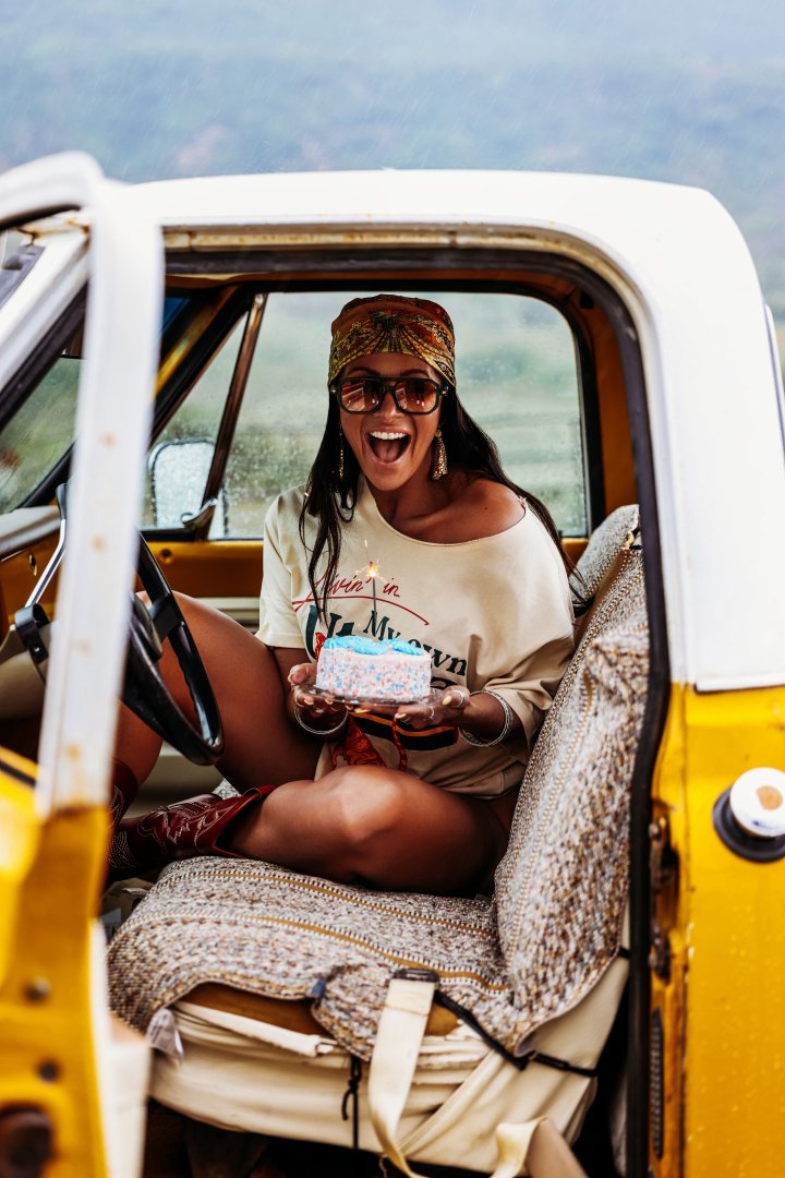 Woman with long dark hair, wearing glasses, a colorful headscarf, and a beige t-shirt, sitting in the driver's seat of a vintage yellow and white vehicle, holding a birthday cake with a lit candle, smiling joyfully.
