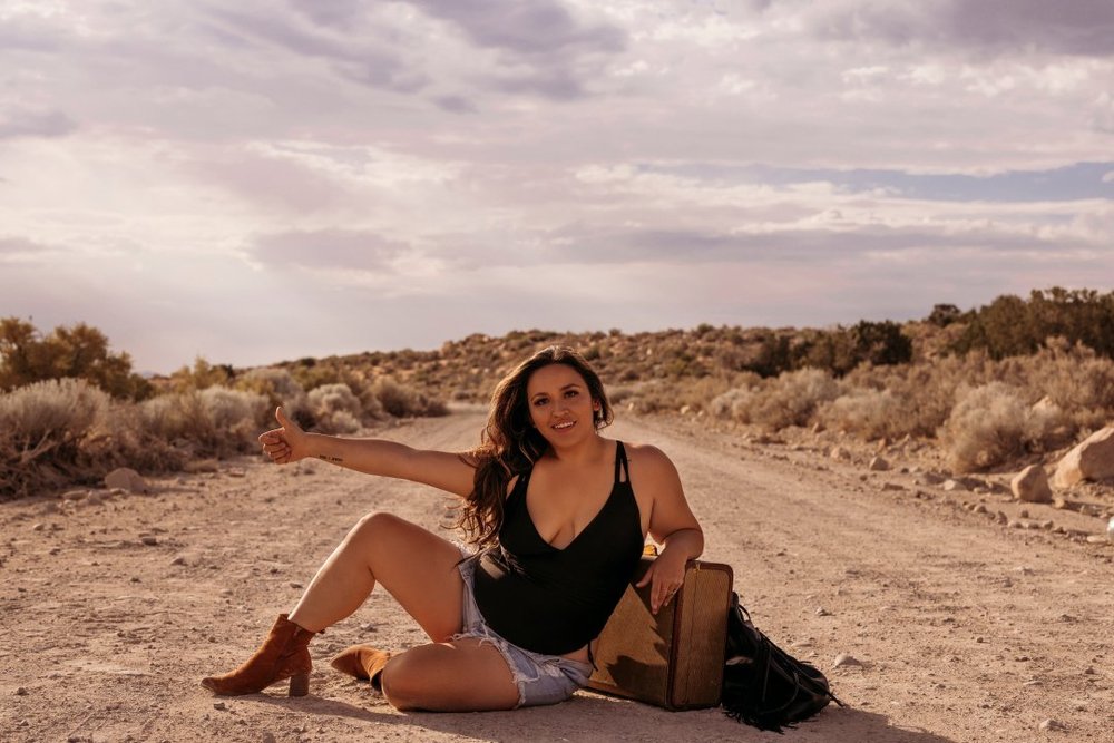 A woman sitting on a desert road, smiling and giving a thumbs-up beside her luggage