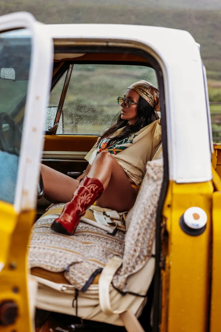 A woman wearing glasses, a headscarf, and a loose beige shirt, sitting in the driver's seat of a yellow and white vintage truck, with her legs crossed and wearing red cowboy boots, in a rural outdoor setting.