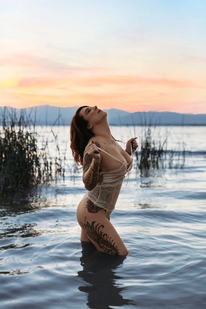 A woman with tattoos standing waist-deep in water at sunset, wearing a lace top and holding her hair with both hands.