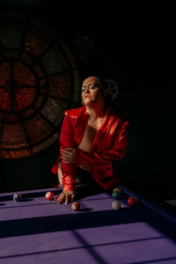 A woman in a red blazer leans over a purple pool table with billiard balls, with a large, colorful clock on a dark background behind her.