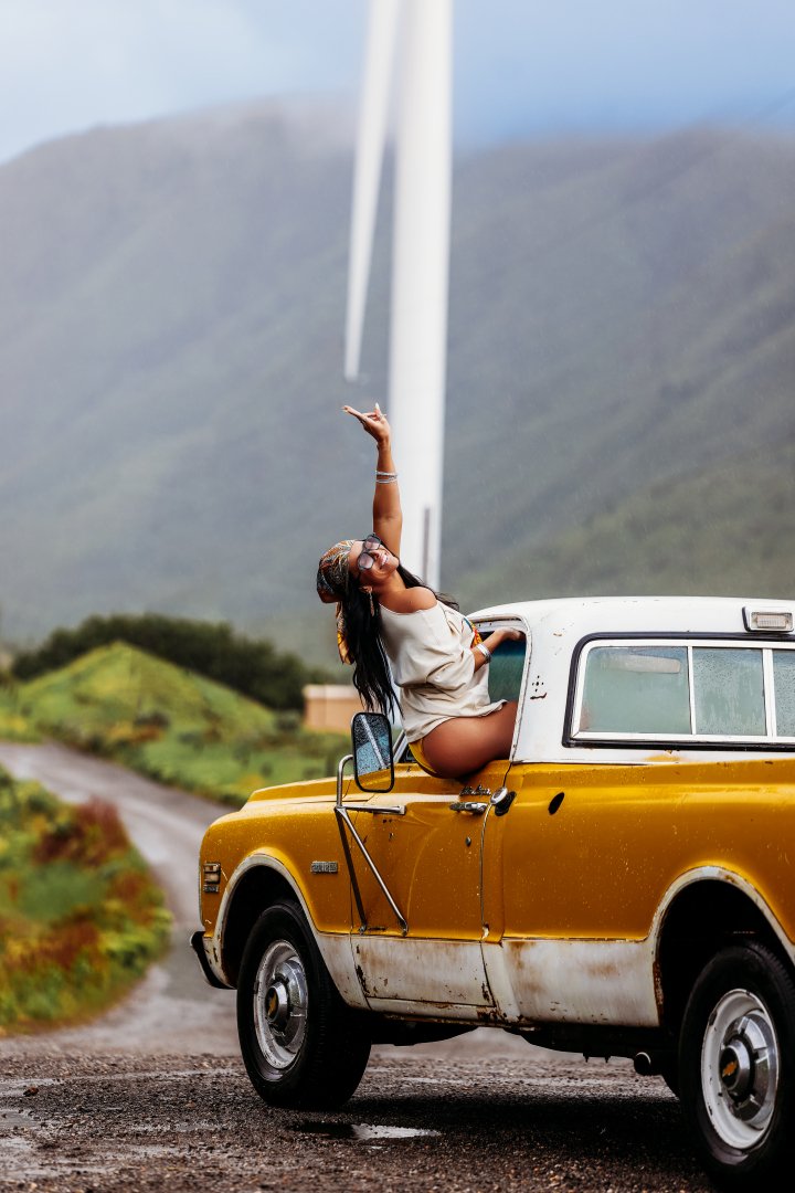 A woman sitting on the edge of a vintage yellow truck with a scenic mountain landscape and wind turbines in the background.