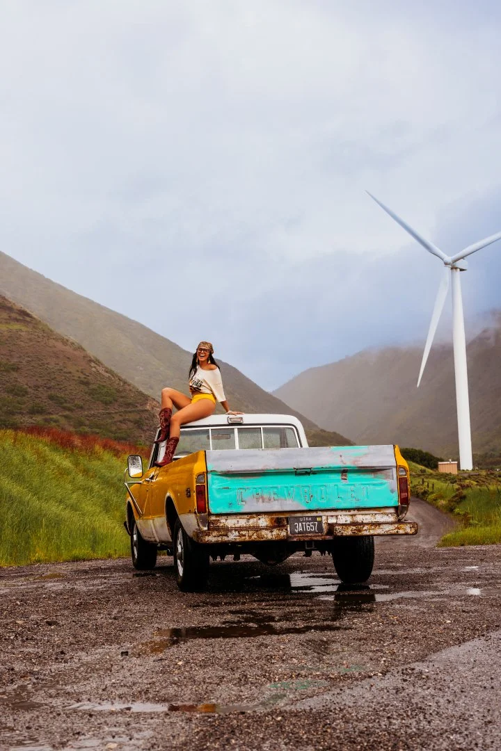 A woman sits on the back of a yellow pickup truck with a weathered tailgate, parked on a rural dirt road. The landscape features green hills, a wind turbine in the distance, and a cloudy sky.