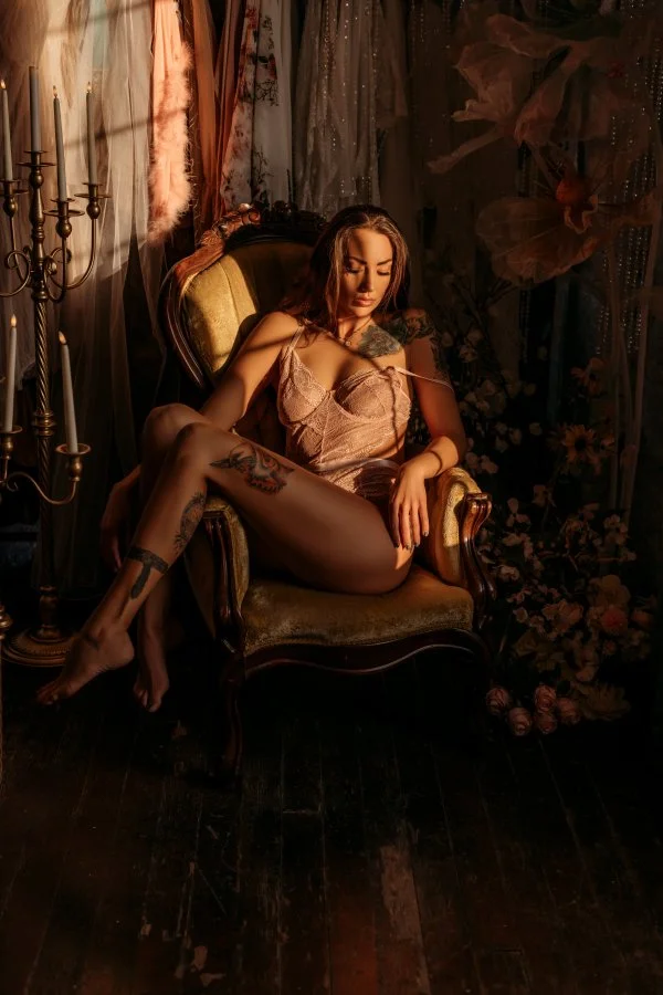 A woman with tattoos sitting on an antique armchair in a dimly lit, floral decorated room wearing lingerie.