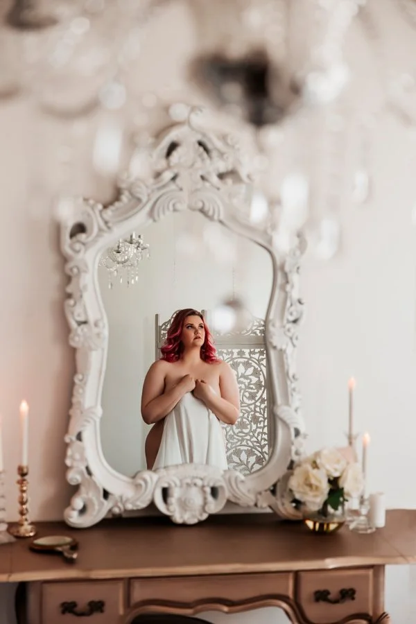 Red-haired breast cancer survivor reflected in an ornate mirror, draped in white sheets with candles and roses