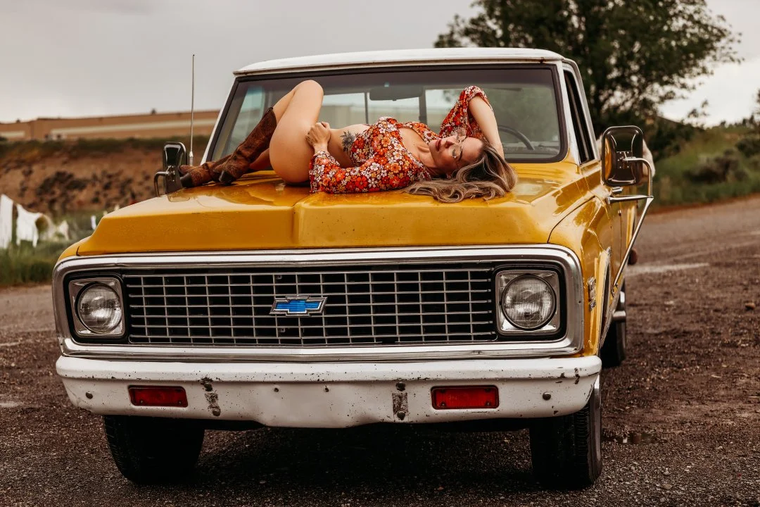 A woman lying on the hood of a yellow vintage Chevrolet truck, outdoors on a dirt road, with grass, trees, and cloudy sky in the background.