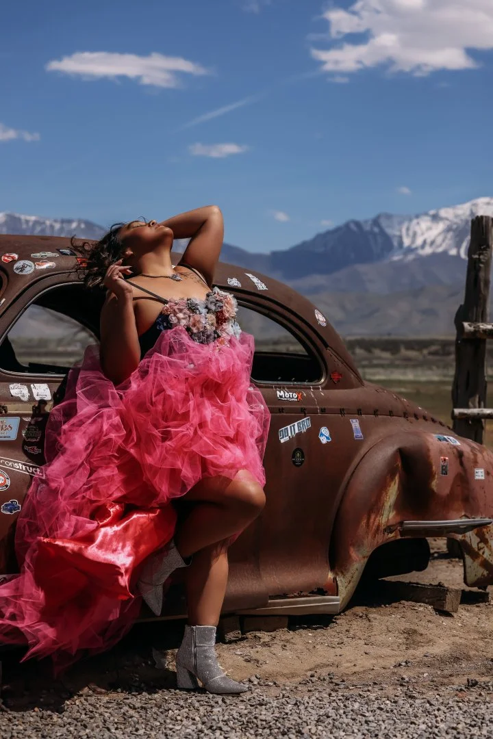 A woman in a pink tulle dress and silver high heels posing in front of an old, rusted car against a mountainous landscape with blue sky and clouds.