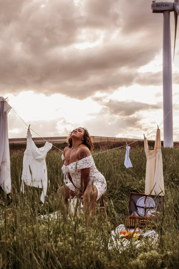 A woman kneeling in a grassy field with a cloudy sky overhead, surrounded by hanging white dresses and a picnic basket on the ground, enjoying the outdoor scene.