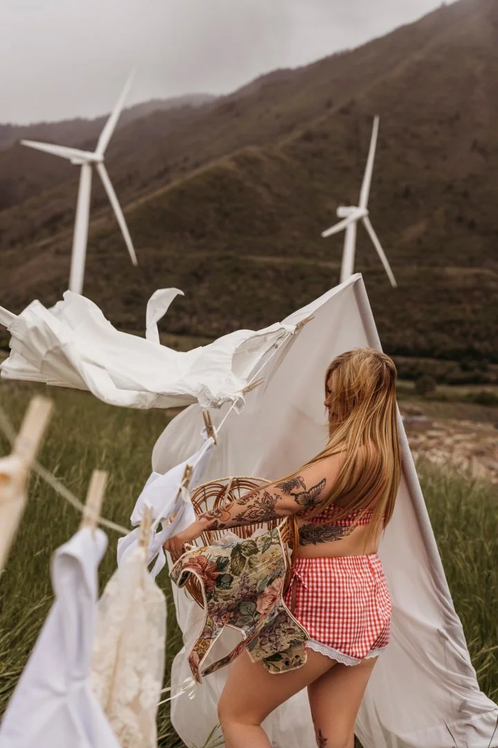 A woman with tattoos on her right arm and long blonde hair, dressed in red and white checkered shorts and a floral shirt, setting up a clothesline with white laundry outdoors in a grassy field with wind turbines on a hill in the background.