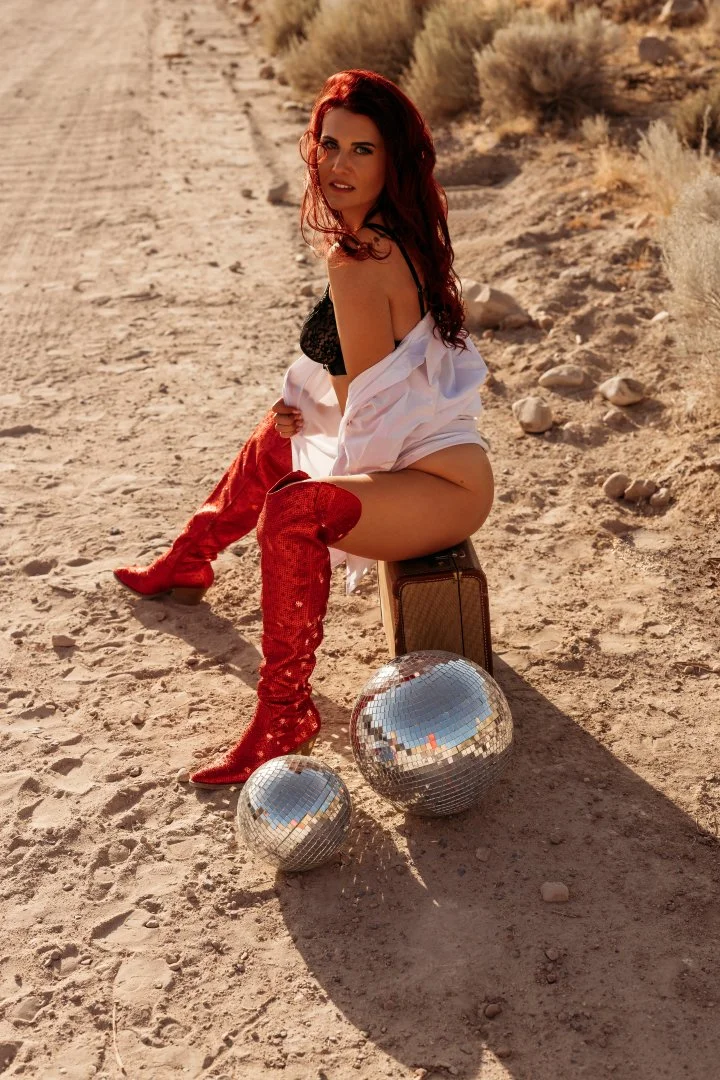 A woman with red hair wearing red boots, black lingerie, and an open white shirt, sitting on a suitcase in a desert setting, with a disco ball and a smaller mirror ball on the ground.