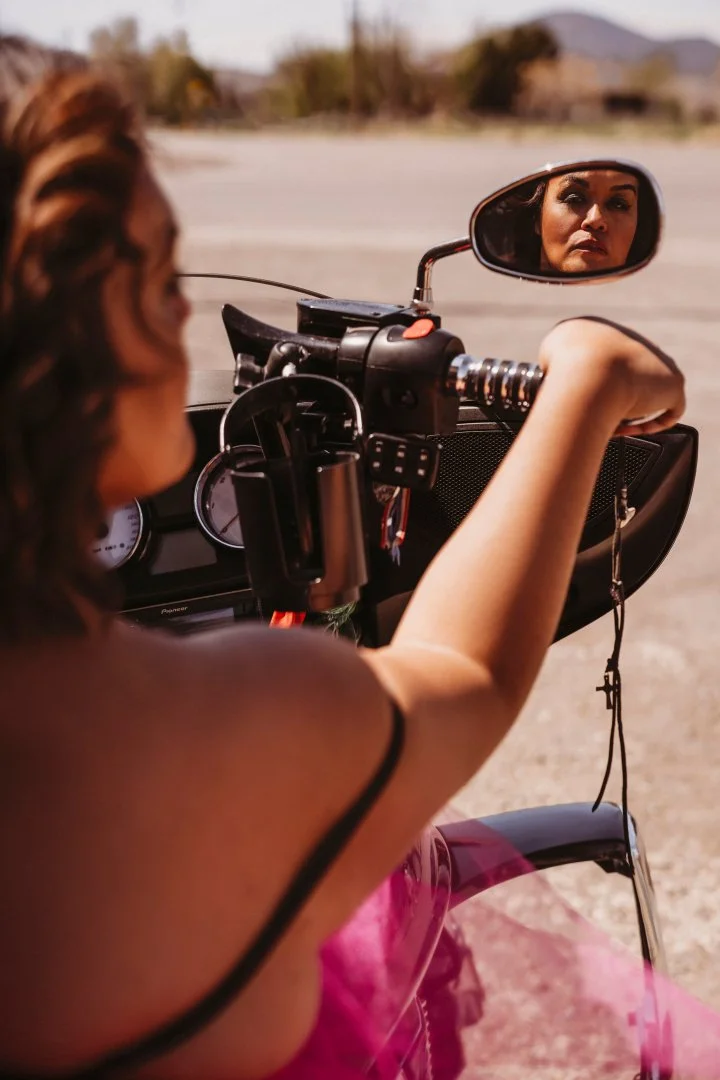 A woman with brown hair sitting on a motorcycle, looking into the rearview mirror, with a blurred outdoor landscape in the background.