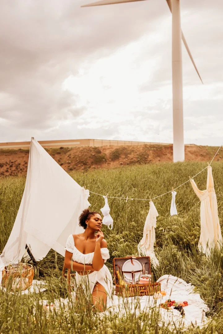 A woman having a picnic outdoors among tall grass with a wind turbine in the background, sitting in front of a picnic basket and surrounded by hanging clothes.