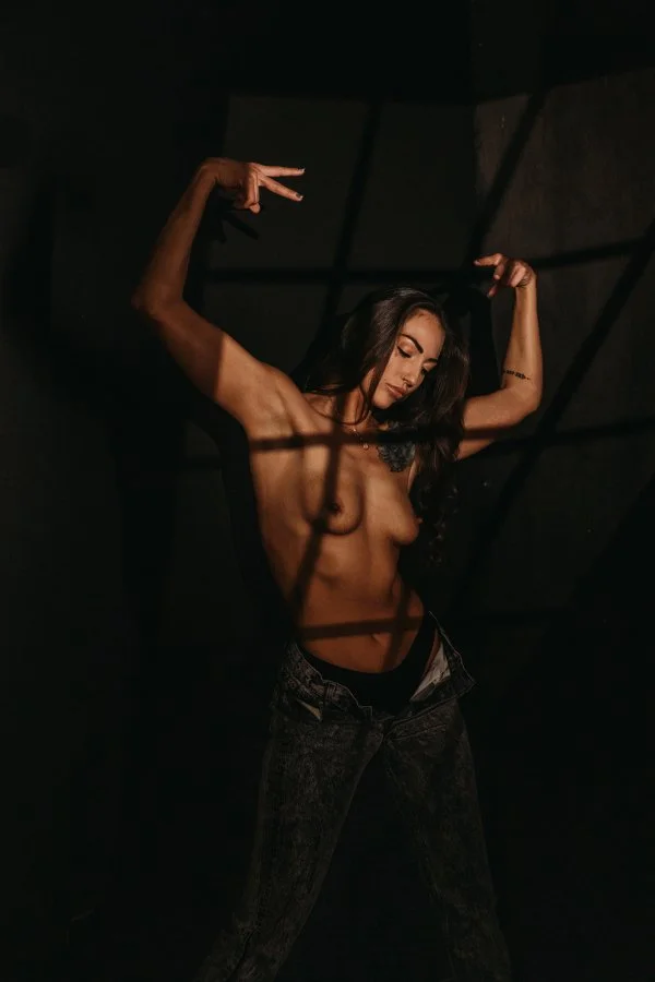 A topless woman with long dark hair poses with her arms raised, making peace signs in a dark setting with dramatic shadows, wearing dark jeans.