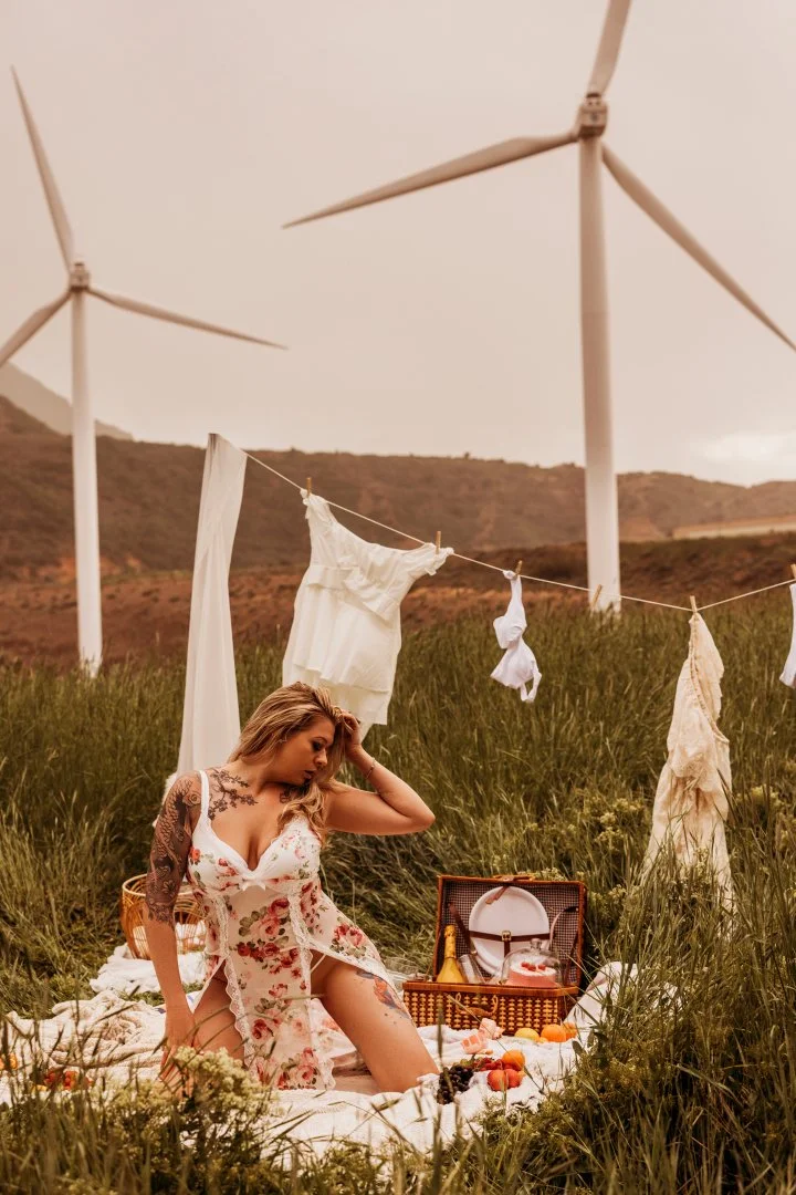 A woman with tattoos in a floral dress sitting on a picnic blanket in a grassy field, with a wind turbine in the background. She is near an open picnic basket and clothes hanging on a line.