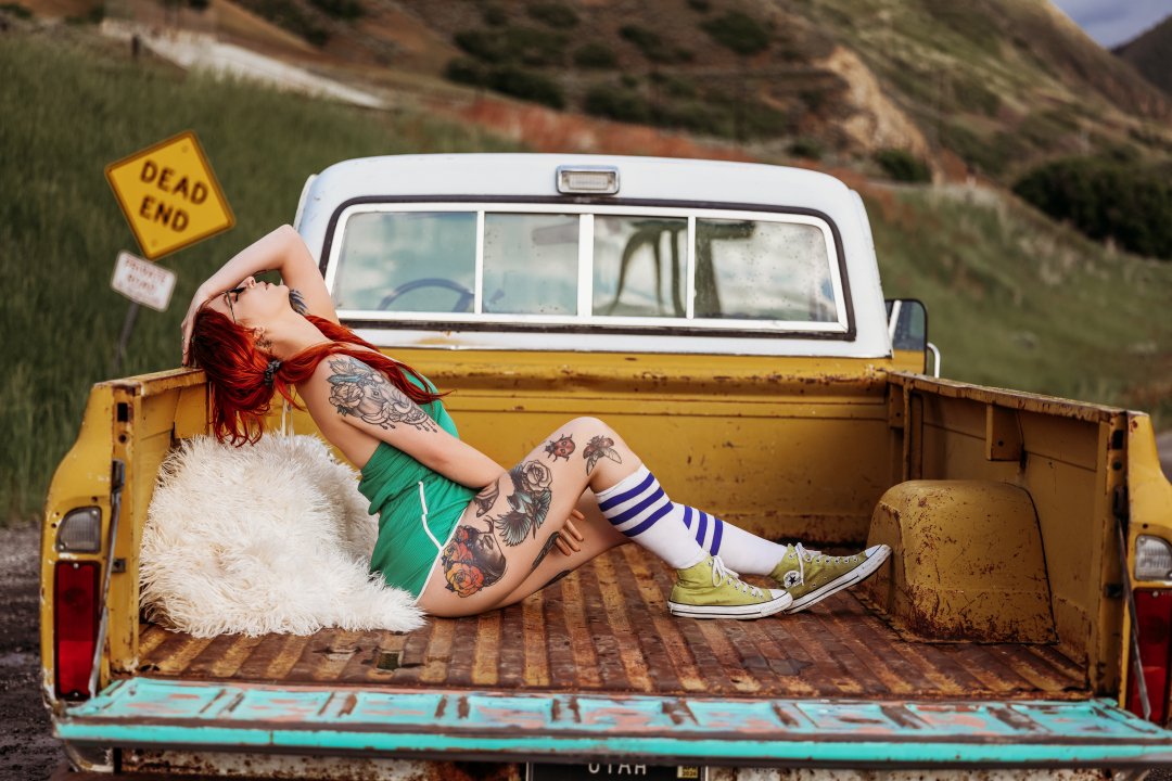A woman with red hair and tattoos, wearing a green tank top, white shorts, striped socks, and yellow shoes, lounging in the back of a yellow pickup truck with a white camper shell, on a rural road with haystacks and hills in the background.
