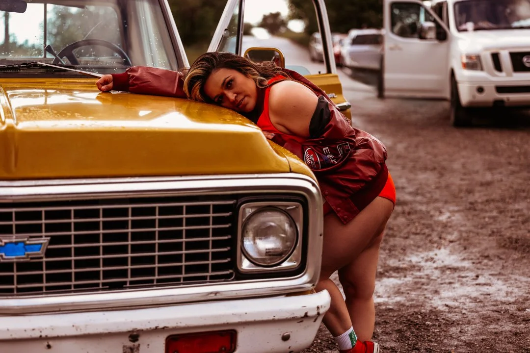 A woman wearing a red dress and a maroon jacket leaning on the hood of a yellow vintage Chevrolet car, looking at the camera with her head resting on her arm. There are other cars in the background on a dirt parking area.